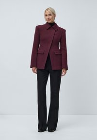 Woman standing wearing a dark plum tailored jacket with a collar and black wide-leg pants against a plain background.