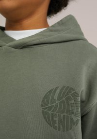Maslinasto zeleni dukserica s kapuljačom izrađena od pamuka, s teksturiranim kružnim logotipom u tamnije zelenoj boji sprijeda. Mekani, udobni materijal s sportskim dizajnom.