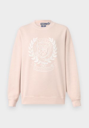 Kvinde iført en lyserød sweatshirt over en stribet skjorte, blå vide jeans, sorte sko, der holder en hvid håndtaske med en guldlås.