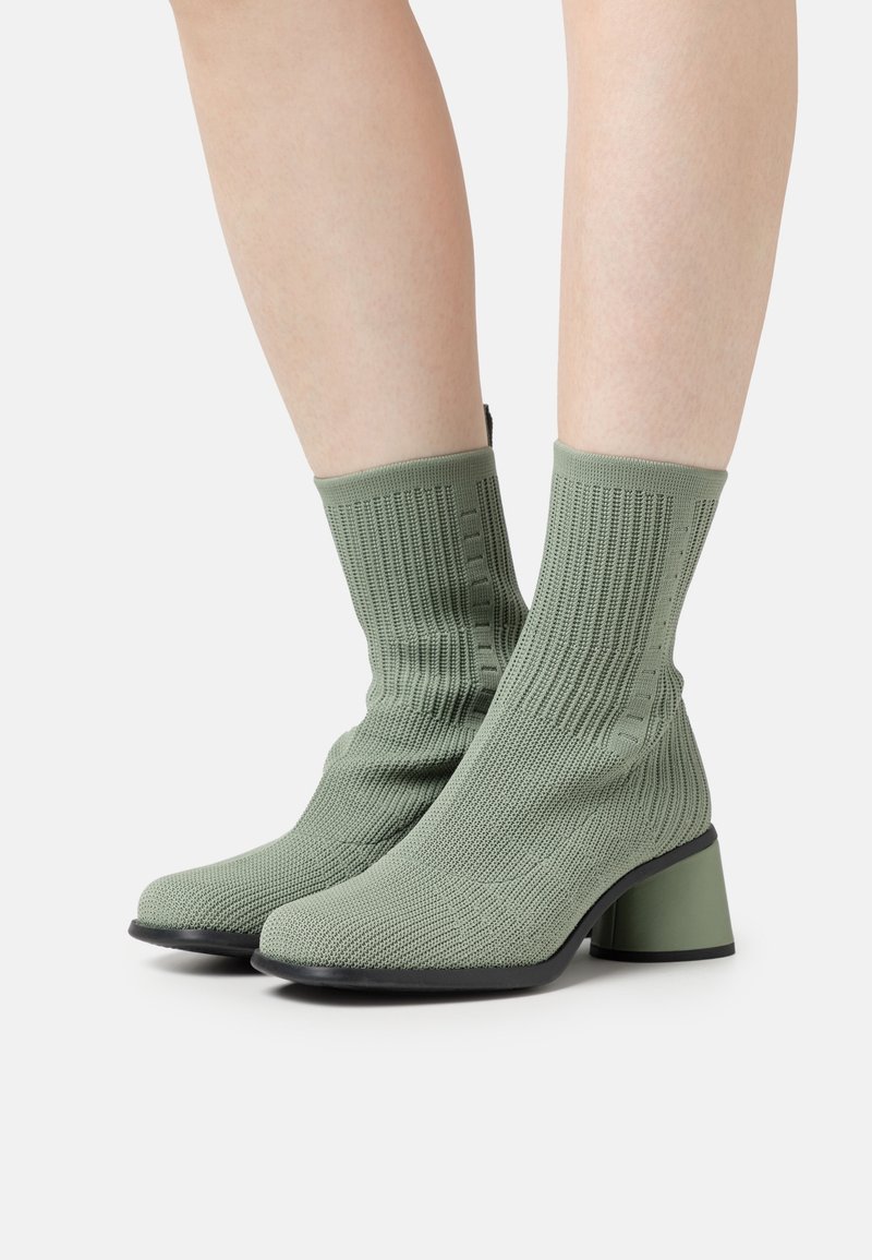 Paire de bottines chaussettes tricotées vert clair à talons carrés portées sur des jambes nues sur fond blanc.