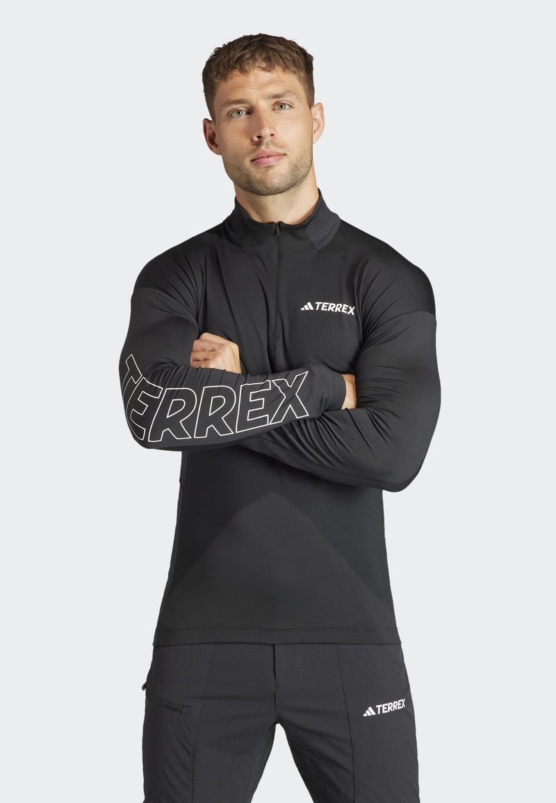 Maglia atletica nera a maniche lunghe con colletto zip. Presenta il logo "TERREX" in bianco sul petto e in grande sulla manica. Tessuto liscio e aderente.