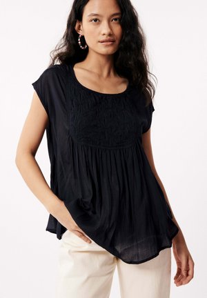 Navy blue short-sleeve blouse with textured embroidered panel, gathered waist, and side pockets, paired with light-colored pants.