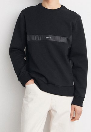 Osoba ubrana w czarną, długorękawową bluzę z poziomym błyszczącym paskiem i napisem "BOSS" na piersi, połączoną z jasnobeżowymi spodniami.