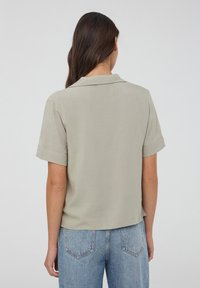 Blusa corta grigia chiaro a maniche corte con colletto a punta e maniche arrotolate, realizzata in un tessuto liscio, abbinata a jeans blu a vita alta.
