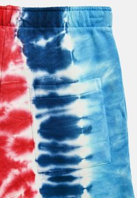 Pantaloni corti tie-dye rossi, bianchi e blu realizzati in tessuto morbido, dotati di una vita elastica e una tasca laterale. I motivi si fondono perfettamente.