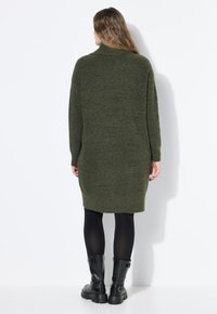 Abito lavorato a maglia verde oliva con collo alto, maniche lunghe e vestibilità ampia, che arriva a metà coscia, abbinato a stivali neri alla caviglia.