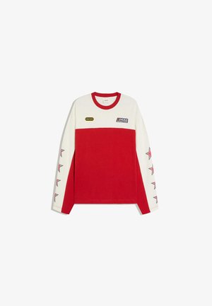 Junger Mann trägt einen rot-weißen Pullover mit Sternenmustern auf den Ärmeln und Text-Patches, steht auf einer Straße in der Stadt in der Nähe von Palmen.