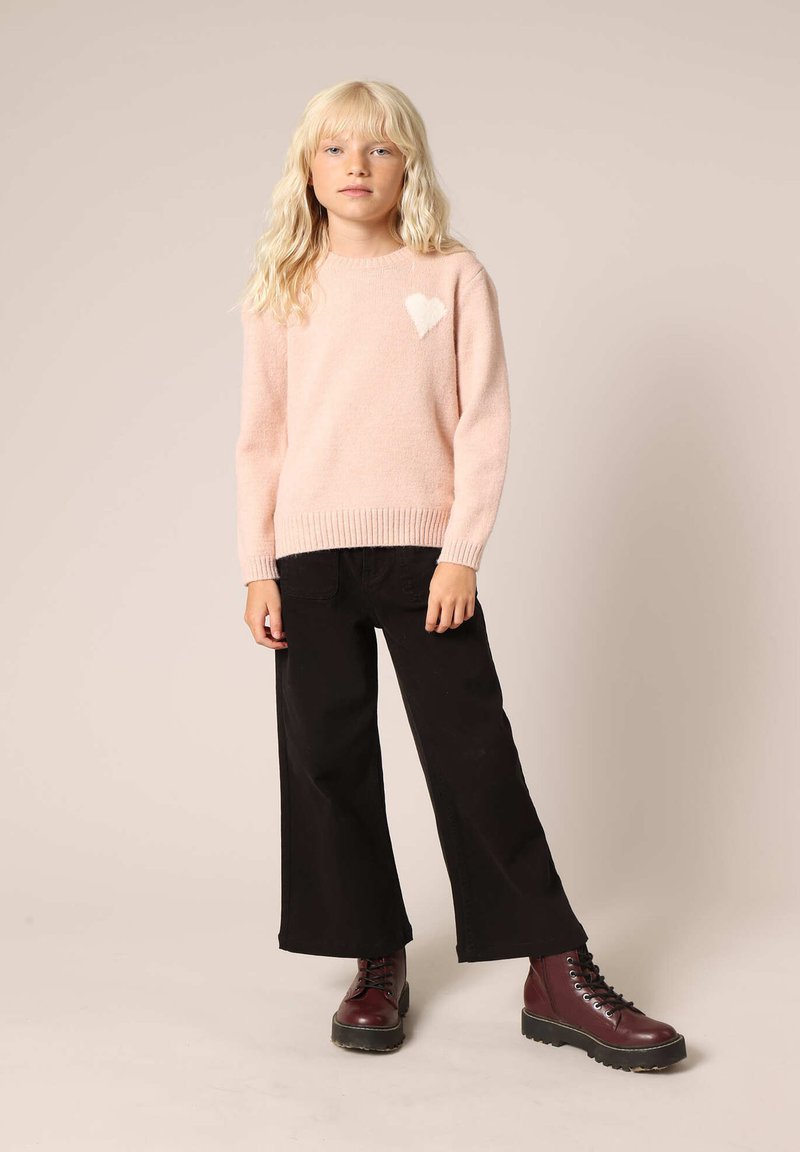 Pull en maille rose clair avec un détail en forme de cœur blanc, associé à un pantalon large noir et des bottes bordeaux à lacets.