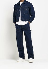 Mörkblå denimjacka med två bröstfickor, tillsammans med matchande löst sittande byxor. Vit t-shirt under med vita sneakers.