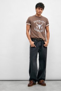 Brun grafisk t-skjorte med design av ku-skalle og kaktus, kombinert med løstsittende svarte jeans og brune snørebåndssko.