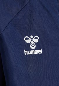 Tissu bleu marine avec un motif texturé. Présente un logo "hummel" blanc avec un design d'abeille stylisé, situé sur le côté gauche.