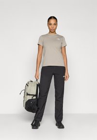 Μπεζ μπλουζάκι με λογότυπο, μαύρο παντελόνι και γκρι σακίδιο με μαύρες λεπτομέρειες. Το μοντέλο στέκεται όρθιο με τα χέρια στα πλάγια σε απλό φόντο.