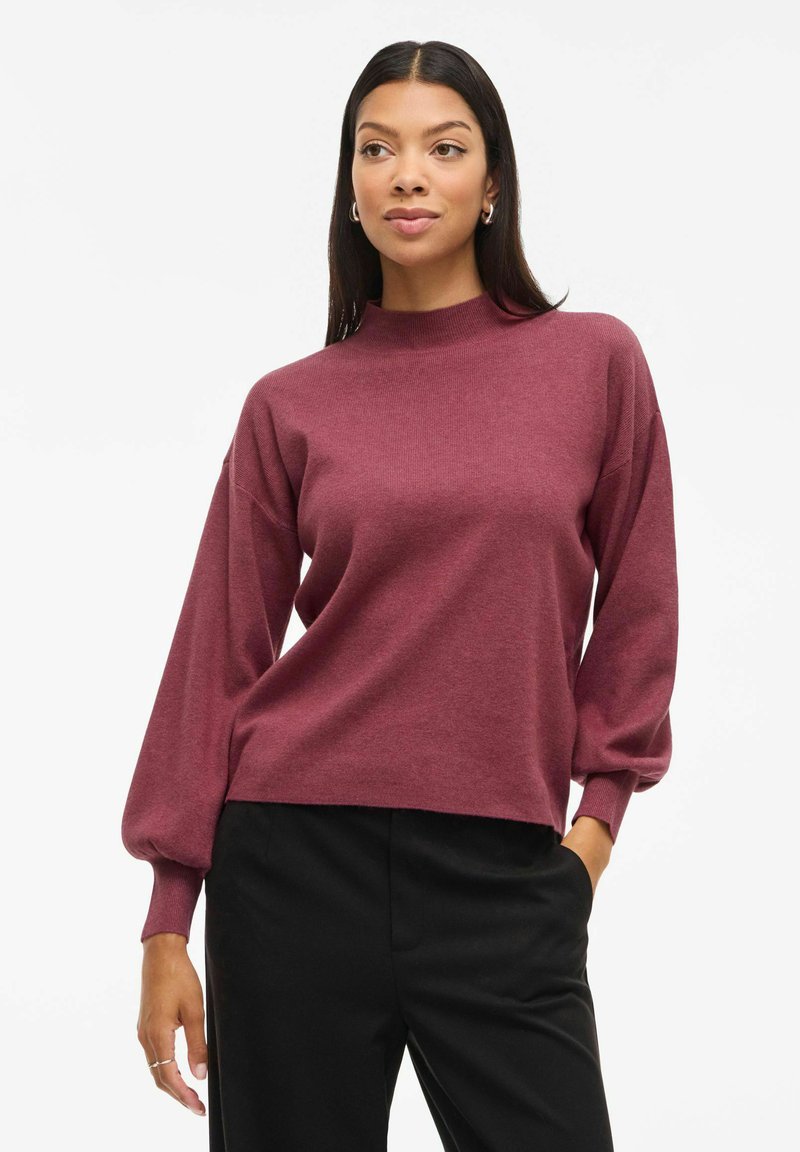 Bordeaux pullover met een hoge ronde hals en luchtige mouwen, gemaakt van zachte stof. Gecombineerd met zwarte broek, wat een relaxte pasvorm laat zien.