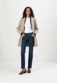 Beige Wollmischung Mantel, weißes eng anliegendes Oberteil, dunkelblaue Jeans und braune Loafers mit goldenen Akzenten. Model steht mit den Händen in den Taschen.