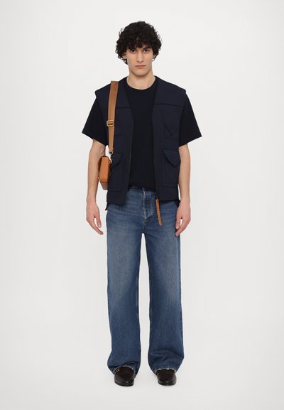 Gilet bleu foncé avec plusieurs poches porté par-dessus un t-shirt noir, associé à un jean large bleu clair et des chaussures marron, avec un sac bandoulière marron.