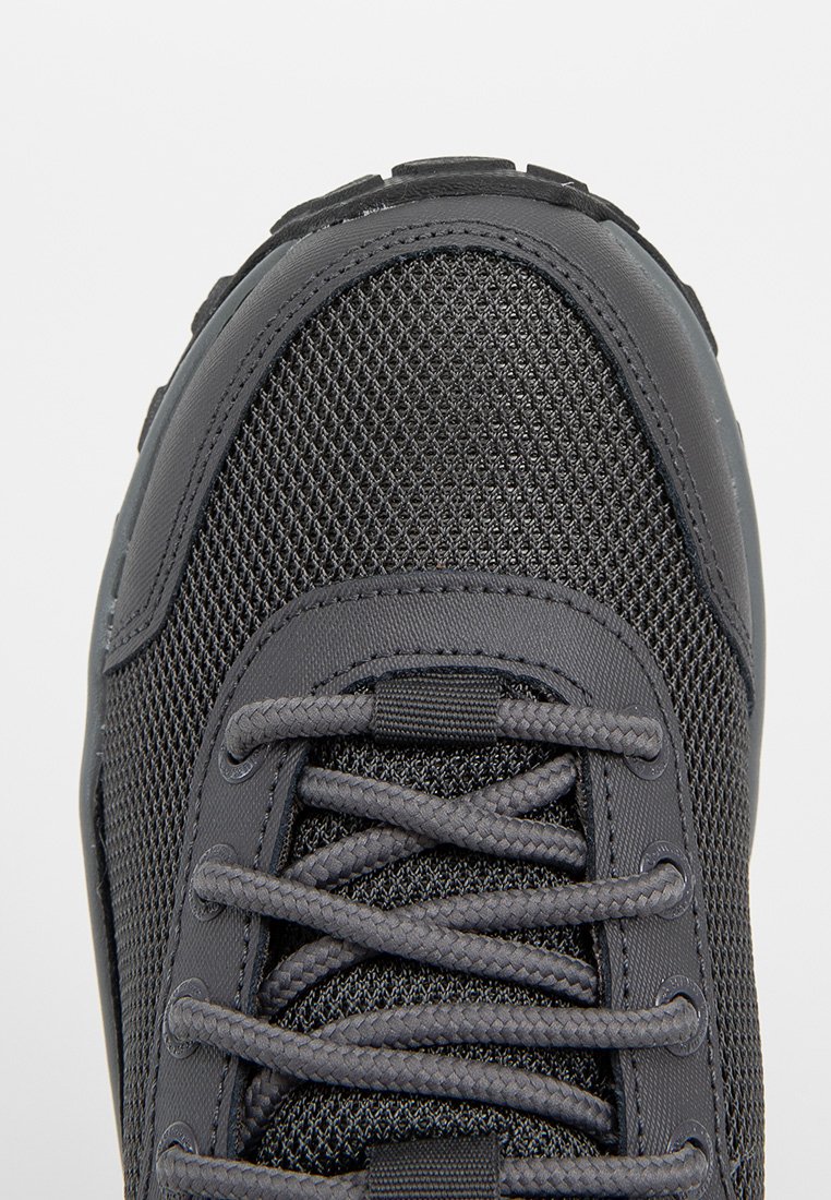 Zapatilla deportiva negra con parte superior de malla texturizada, puntera reforzada y cordones planos. Presenta una suela de goma duradera y un diseño minimalista.