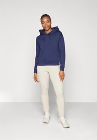 Marineblå bomullshettegenser med frontlomme og justerbar snor i hetten, kombinert med kremfargede slim-fit bukser og hvite joggesko.