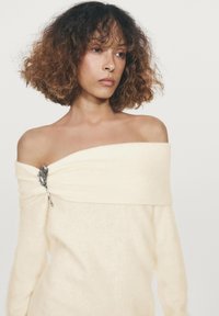 Off-white, off-shoulder sweater med en blød tekstur og draperet design, med en sølvfarvet blomsterbroche accent på venstre skulder.