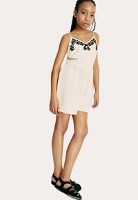 Beige ærmeløs jumpsuit med sort blomsterbroderi langs udskæringen, elastisk talje og løse shorts. Sorte flade sandaler bæres.