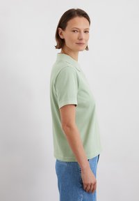 Lichtgroen polo shirt met korte mouwen, kraag en zachte structuur. Gecombineerd met high-waisted blauwe jeans, zij-aanzicht.