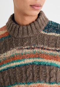 Suéter de lana marrón con un cuello alto con canalé, que presenta franjas horizontales multicolores en naranja, verde azulado y crema, con patrones texturizados.