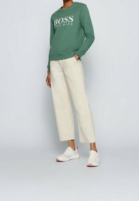 Person som bär en grön Hugo Boss-tröja, off-white vida byxor och vita sneakers står med ena handen i fickan.