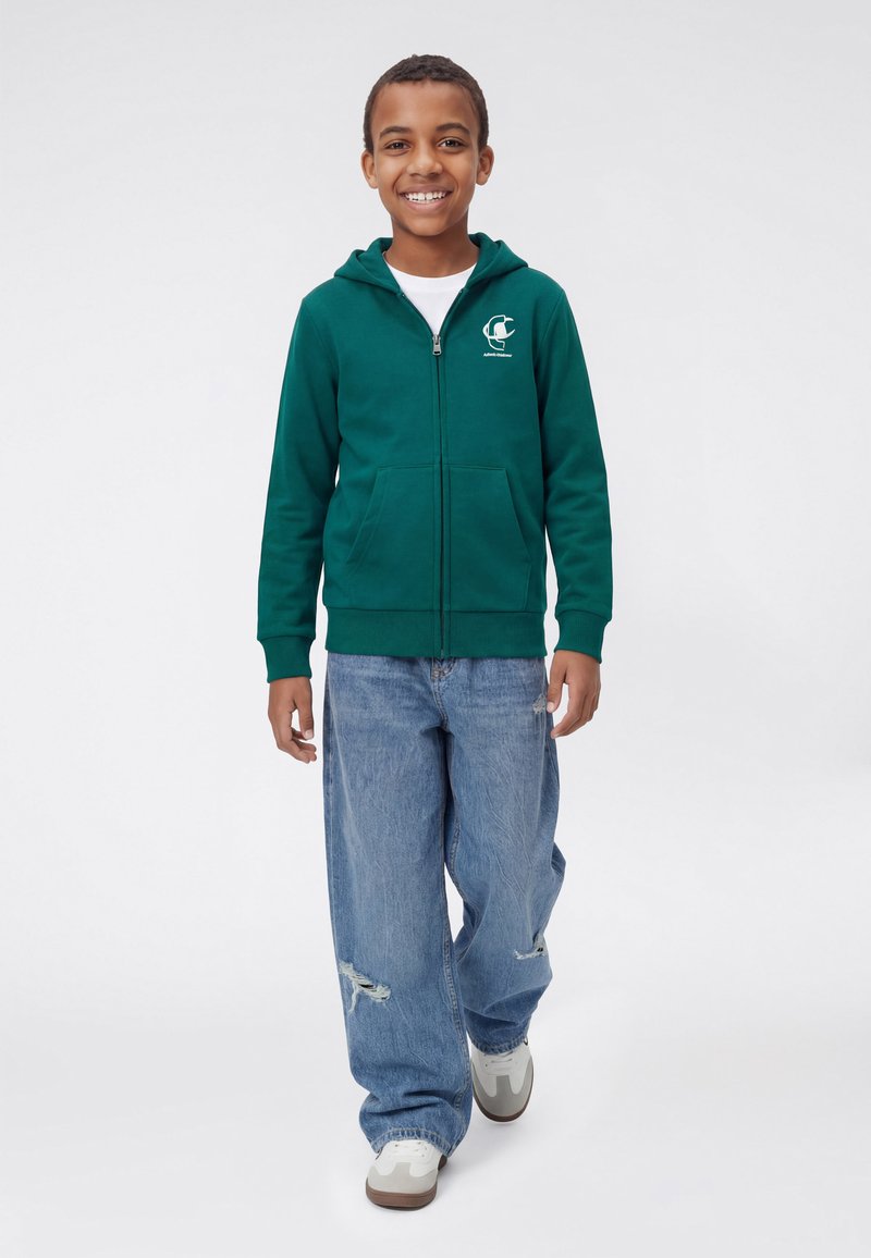 Ragazzo sorridente che cammina avanti indossando una felpa con zip verde, maglietta bianca, jeans blu larghi e strappati e scarpe da ginnastica bianche.