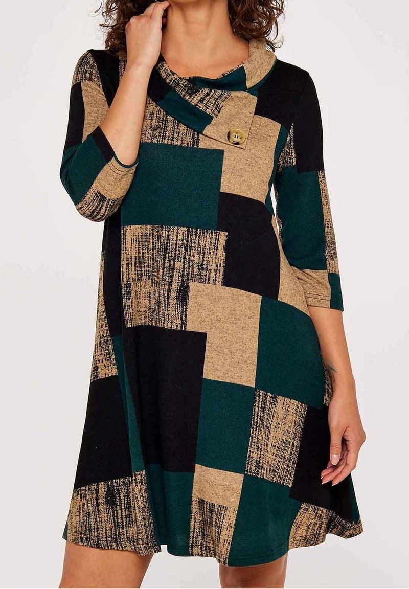 Femme portant une robe mi-longue à manches longues avec un grand motif patchwork vert, noir et beige et un col bénitier boutonné.