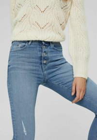 Jeans de tiro alto en azul claro con cinco botones, con un ligero difuminado y un sutil detalle desgastado en la pierna derecha, combinados con un suéter de punto color crema.