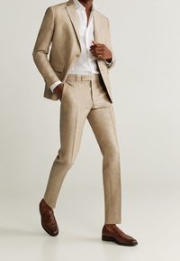 Jasno-beżowy lniany garnitur z dopasowaną marynarką i spodniami, zestawiony z białą koszulą i brązowymi skórzanymi mokasynami. Gładka faktura i doskonale skrojony krój.
