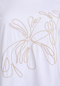 Camiseta blanca con un diseño de bordado texturizado en forma de cuerda beige que se enrosca por la tela. Material de algodón suave.