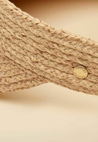 Close-up of woven beige straw visor with braided texture and small round gold logo tag.