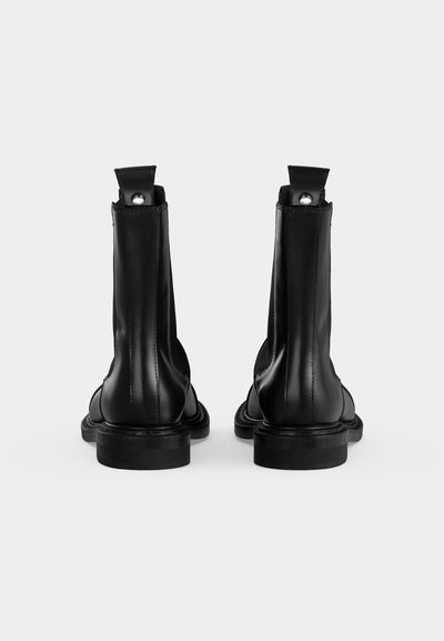 Paire de bottines en cuir noir avec languettes de traction et détails de couture, vues de dos sur un fond clair uni.