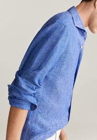 Camisa de lino azul con una textura sutil, cuello abotonado y mangas remangadas. Diseño entallado, dobladillo ligeramente curvado y puños estándar.