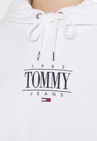 Bílá mikina s kapucí vyrobená z měkké tkaniny, se středovým potiskem "TOMMY" v černé barvě, rokem "1985", "JEANS" pod ním a malým logem v červené, bílé a modré barvě.