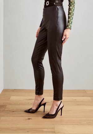 Vrouw draagt zwarte hoog getailleerde nep-lederen broek en zwarte maasjes high-heel slingback schoenen, staand op een houten vloer.