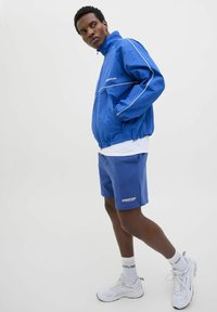 Plavi windbreaker s bijelim detaljima, uparen s plavim šortsem. Model nosi bijele tenisice i kratke čarape, pokazujući ležeran sportski stil.