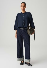 Marineblaues Blusenshirt mit Rundkragen, lockerer Passform, kombiniert mit dunklen, weit geschnittenen Jeans mit umgeschlagenen Säumen und einer geflochtenen Tasche.