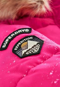 Žiarivo ružová zateplená bunda s vodnými kvapkami, so vyšitými nášivkami: "SUPERDRY®" a "EXPEDITION EVEREST", ktoré zobrazujú hory a slnko.