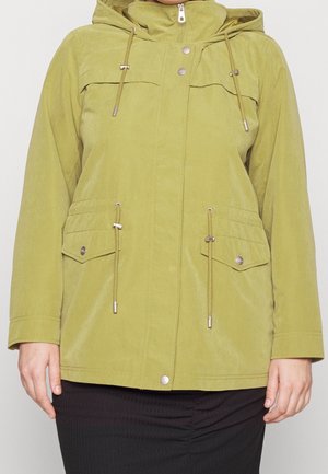 Veste à capuche verte avec fermeture éclair devant, boutons-pression, cordons réglables et poches à rabat portée sur une jupe noire.
