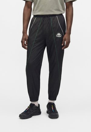Homme portant un pantalon de survêtement noir Kappa avec des détails de bande verte et blanche, des chaussettes blanches et des baskets noires avec des accents colorés.