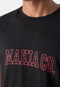 Homme portant un t-shirt noir à col rond avec le texte « MAKIA CO. » en contour rouge sur la poitrine.