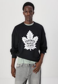 Hanorac negru cu grafică albă "Toronto Maple Leafs", având un design mare cu o frunză. Purtați peste un tricou verde deschis și asortat cu blugi gri.
