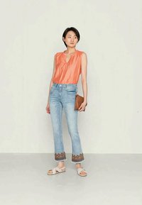 Top laranja sem mangas combinado com jeans claros de boca de sino, apresentando detalhes bordados na bainha. O modelo segura uma clutch castanha e usa sandálias.