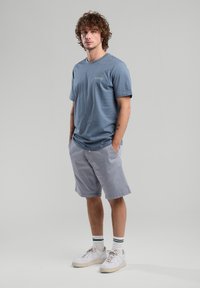 Blue cotton t-shirt with short sleeves and a small chest logo, paired with light gray shorts, standing on a neutral background. White sneakers with green accents.