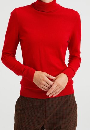 Personne portant un pull à col roulé rouge vif et un pantalon marron à carreaux, les mains doucement jointes devant.