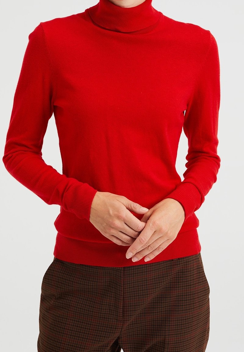Personne portant un pull à col roulé rouge vif et un pantalon marron à carreaux, les mains doucement jointes devant.