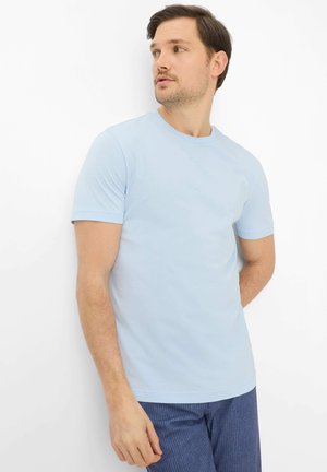 Mand iført lyseblå kortærmet t-shirt og blå bukser med striber, stående mod en hvid baggrund, kigger til siden.