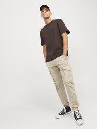 Tricou din bumbac maro, croială lejeră, cu mâneci scurte. Pantaloni cargo bej cu buzunare laterale, picior îngustat, accentuați de adidași negri cu guler înalt.