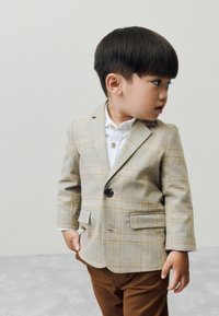 Un jeune enfant portant un blazer à carreaux et une chemise blanche se tient de côté, regardant pensivement vers la gauche sur un fond neutre.